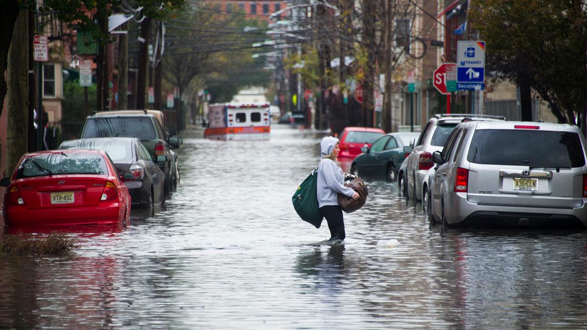 A woman walks through floodwater in the aftermath of Superstorm Sandy in Hoboken, with a stalled ambulance in the background.